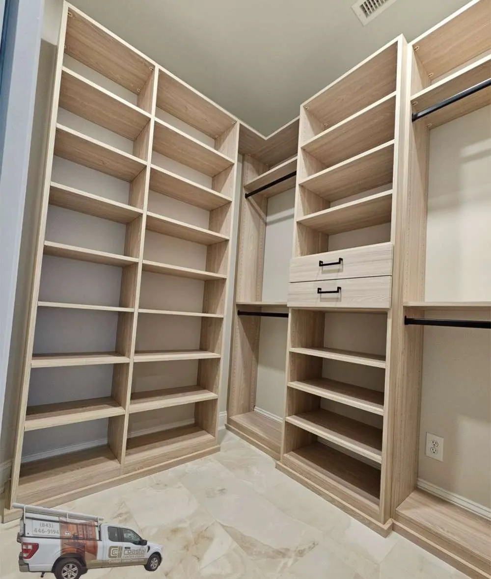 Empty light wood walk in closet with tall corner shelving, two drawers, black hardware, hanging rods and tile floor
