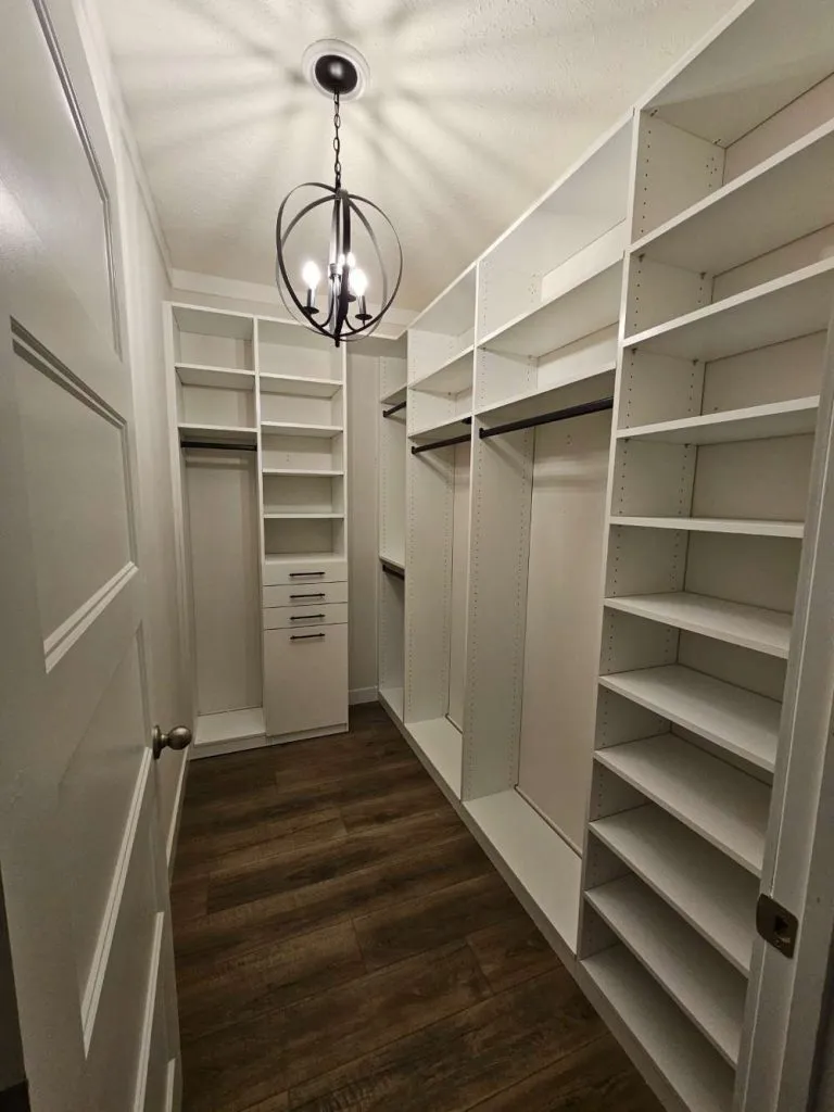 White walk in closet with angled shoe shelves, black hanging rods, orb chandelier and dark wood-look flooring