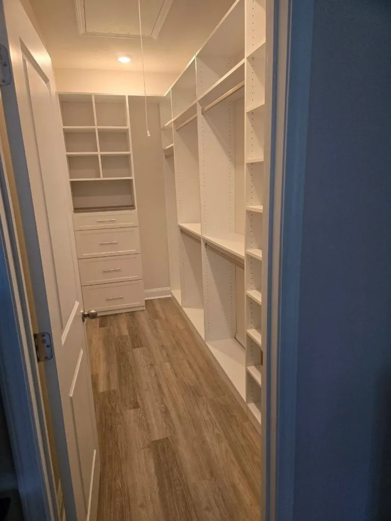 Narrow white walk in closet with tall shelving, hanging rods, a four-drawer unit and wood-look flooring